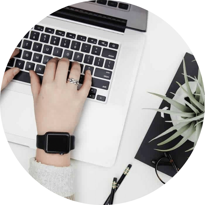 Mains tapant sur un ordinateur portable argenté, avec une montre noire, une bague, une plante verte, deux crayons noirs et un carnet sur fond blanc.